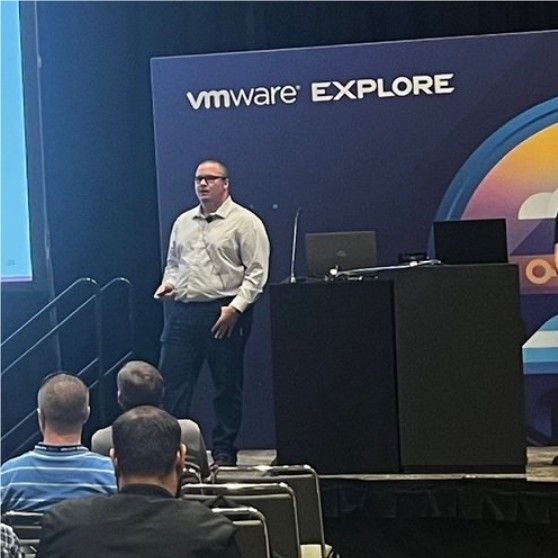 Person presenting at a VMware Explore event, standing on a stage next to a podium with laptops.